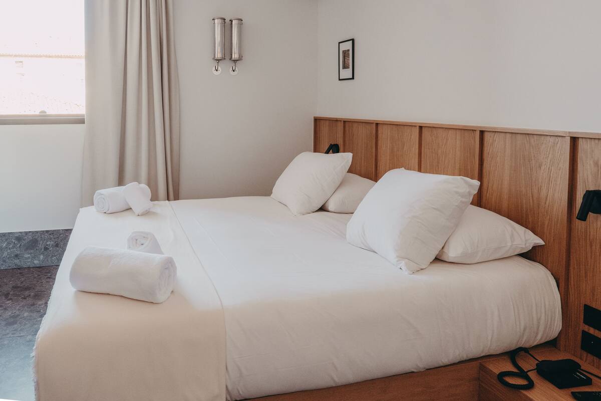 A neatly made bed with white linens and pillows, a wooden headboard, a bedside table with a black phone, and a window with curtains.