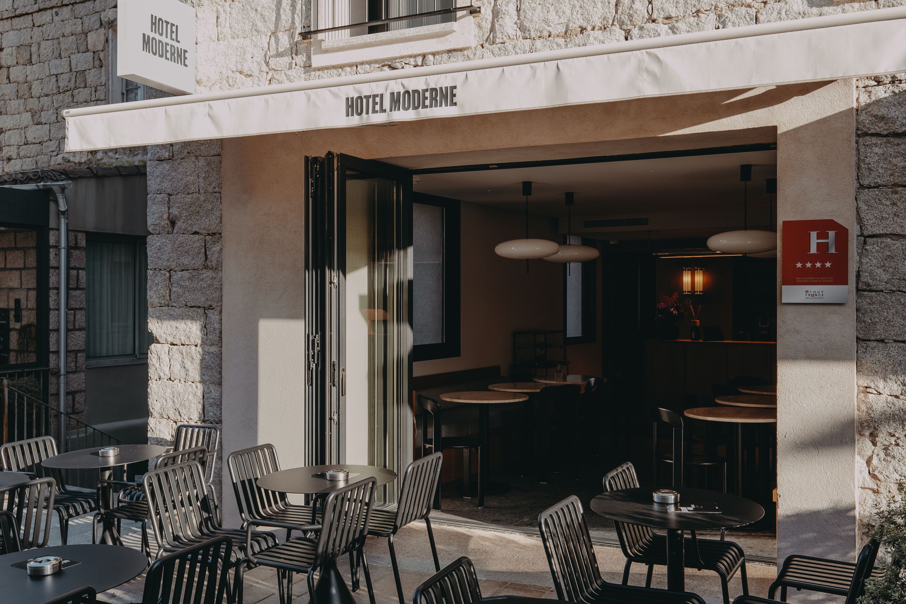 Un espace extérieur aménagé avec des tables et des chaises, une enseigne indiquant « HÔTEL MODERNE », et une vue partielle sur l’intérieur.