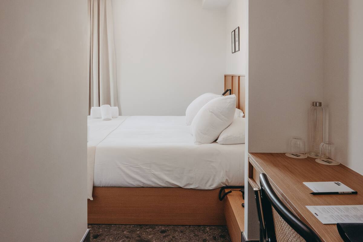 A small, neatly arranged hotel room with a single bed, a wooden desk, and a chair.