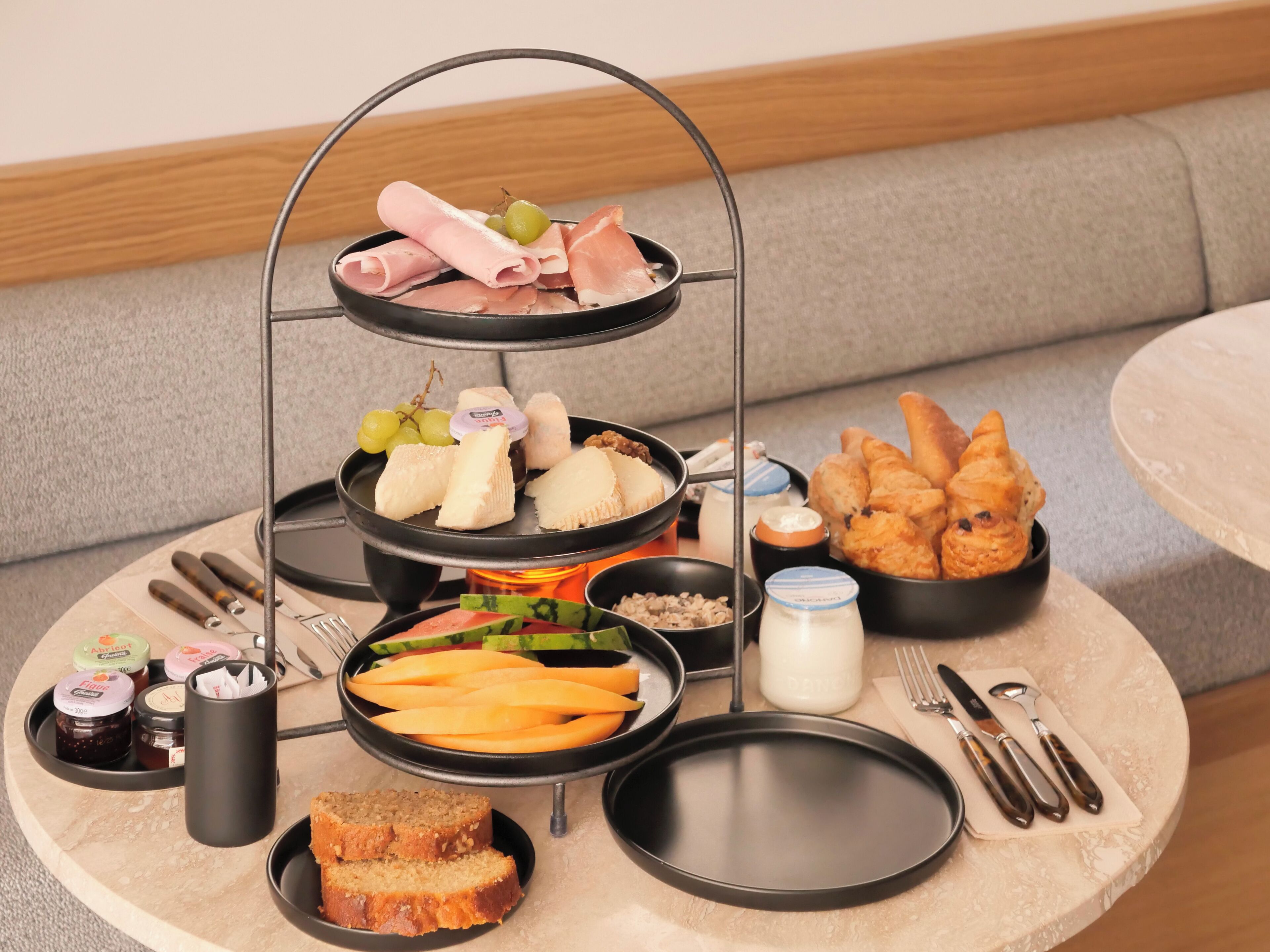 Una bandeja de tres niveles con diversos alimentos, como carnes, quesos y pan, sobre una mesa redonda.