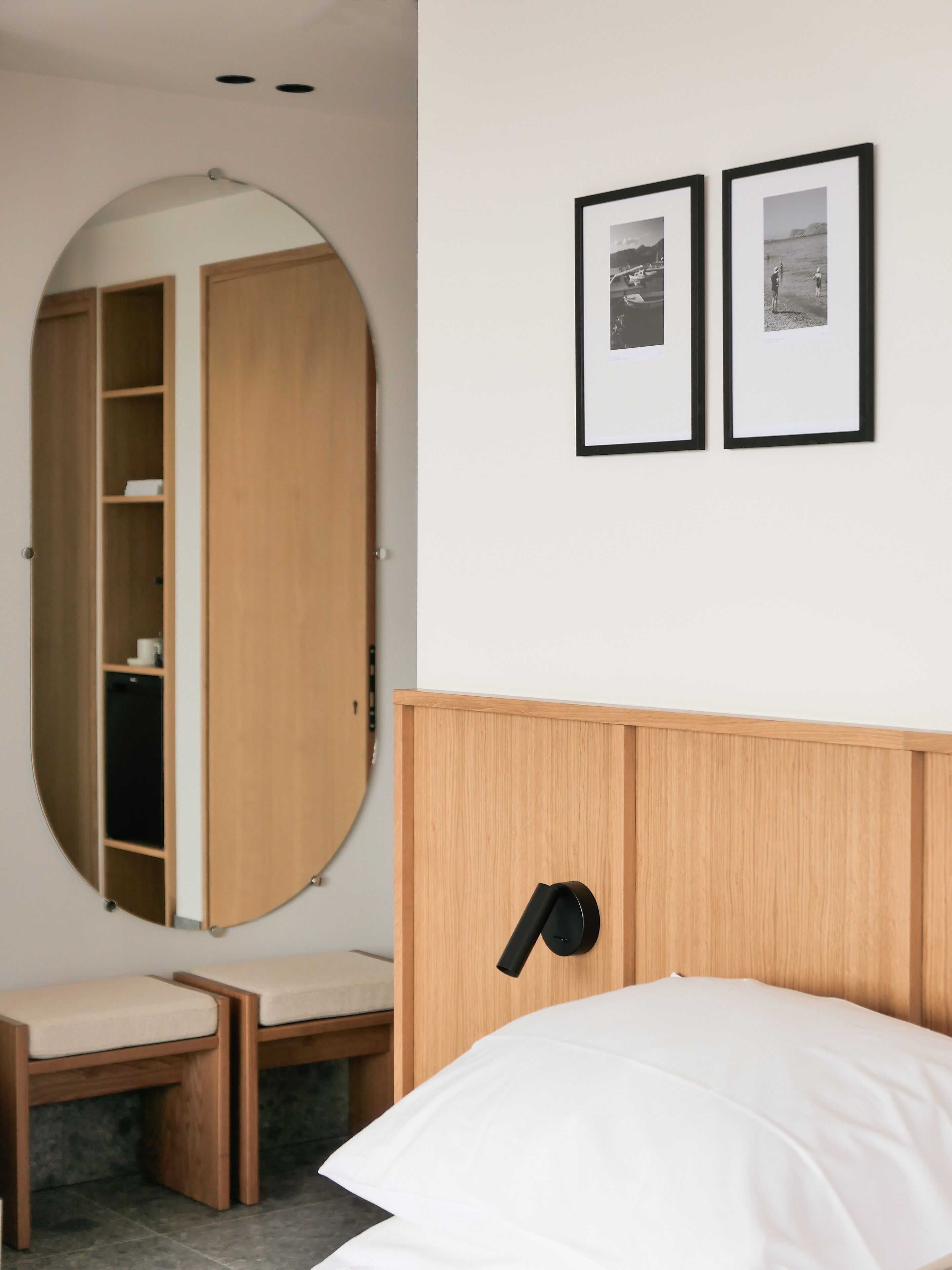 A bedroom with a wooden headboard, a bed with white bedding, a wooden wardrobe, a round mirror, and two framed pictures on the wall.
