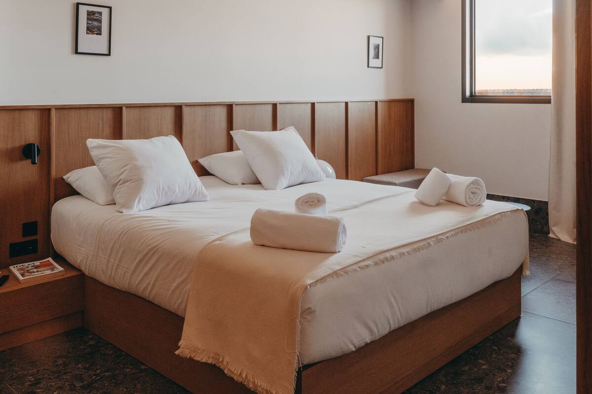A neatly made bed with white linens and a beige throw blanket, a wooden headboard, and a framed picture on the wall.