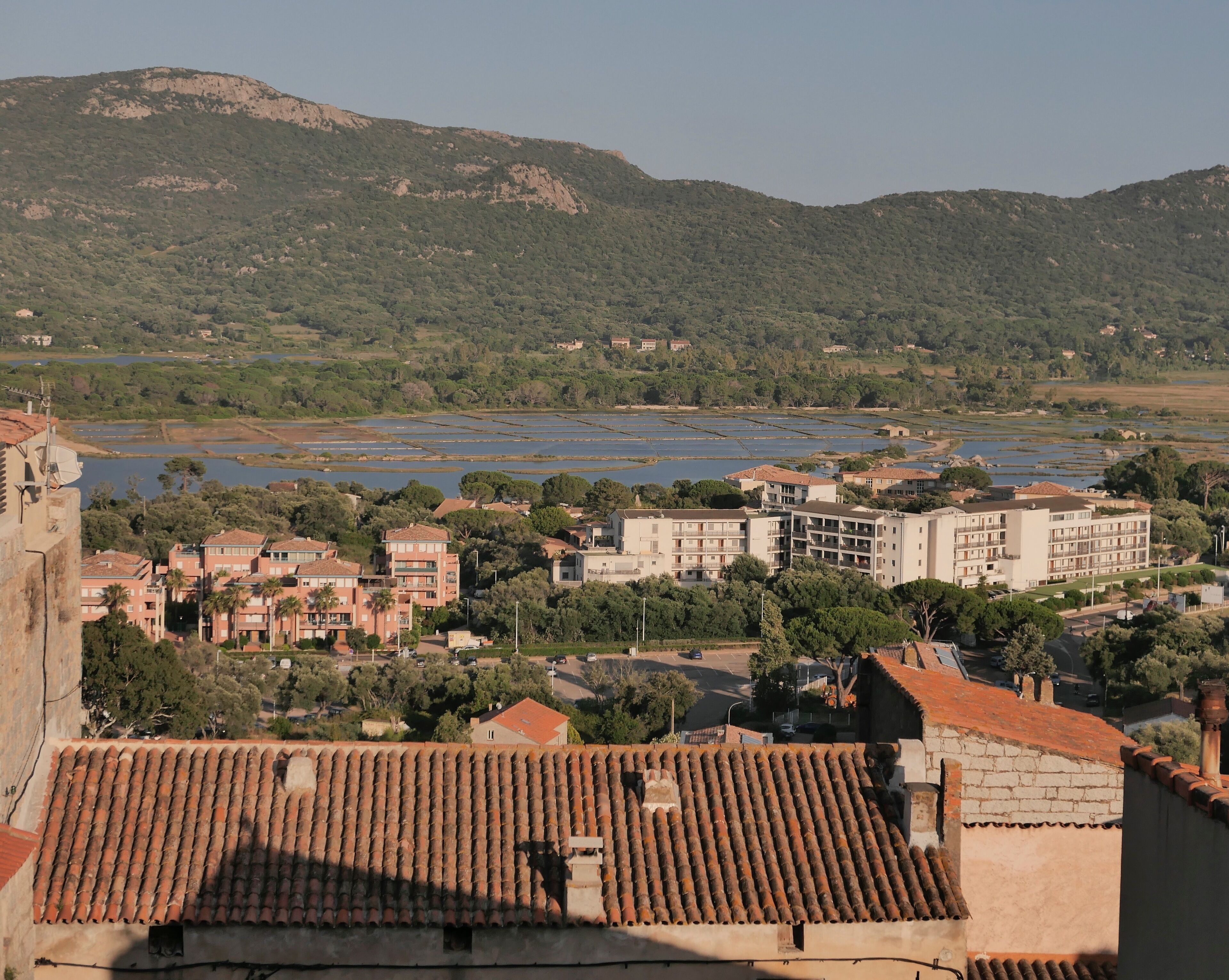 A coastal town with terracotta-roofed buildings, a marina, and a backdrop of hills.