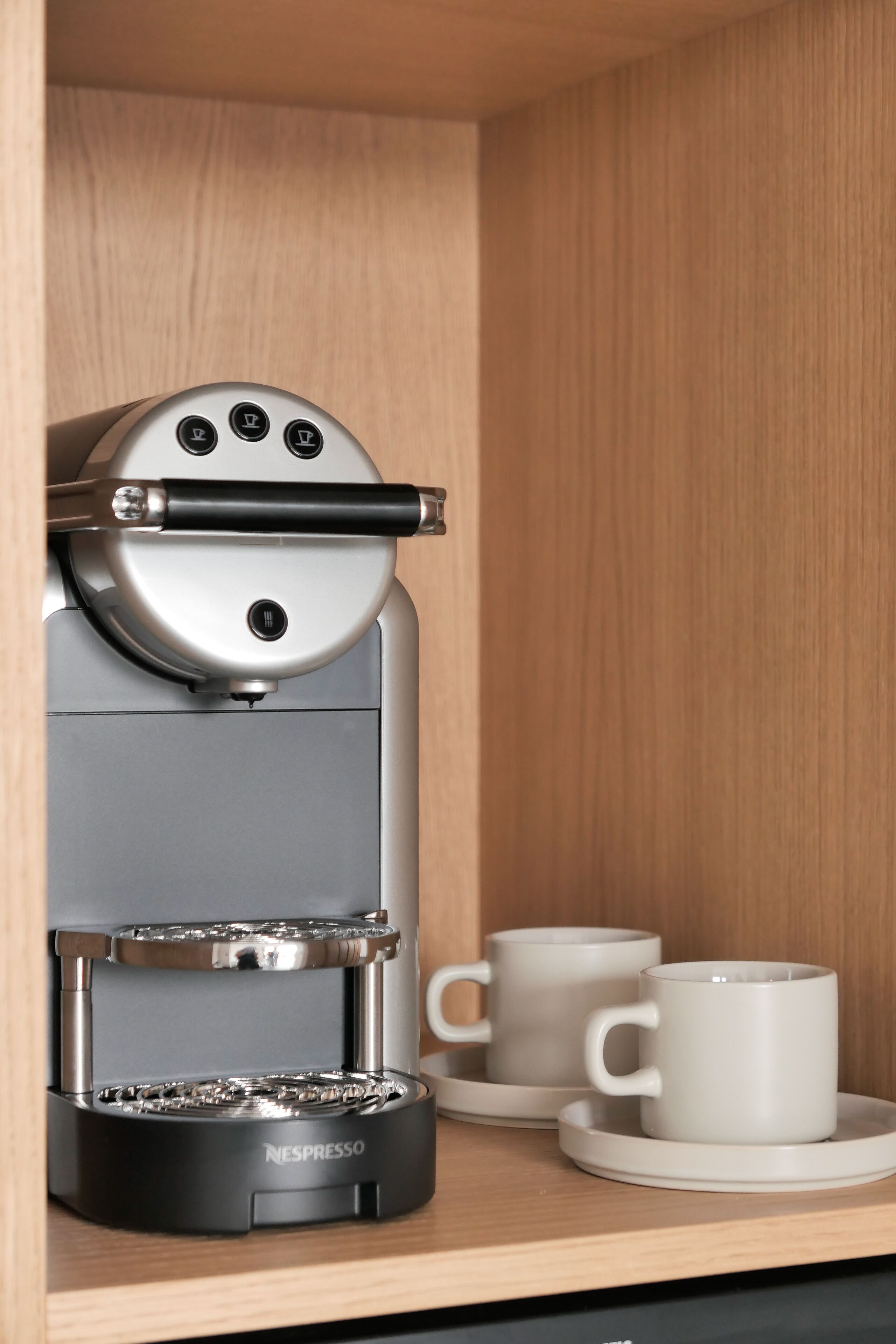 A Nespresso coffee machine in a wooden cabinet with two white cups on a matching saucer.