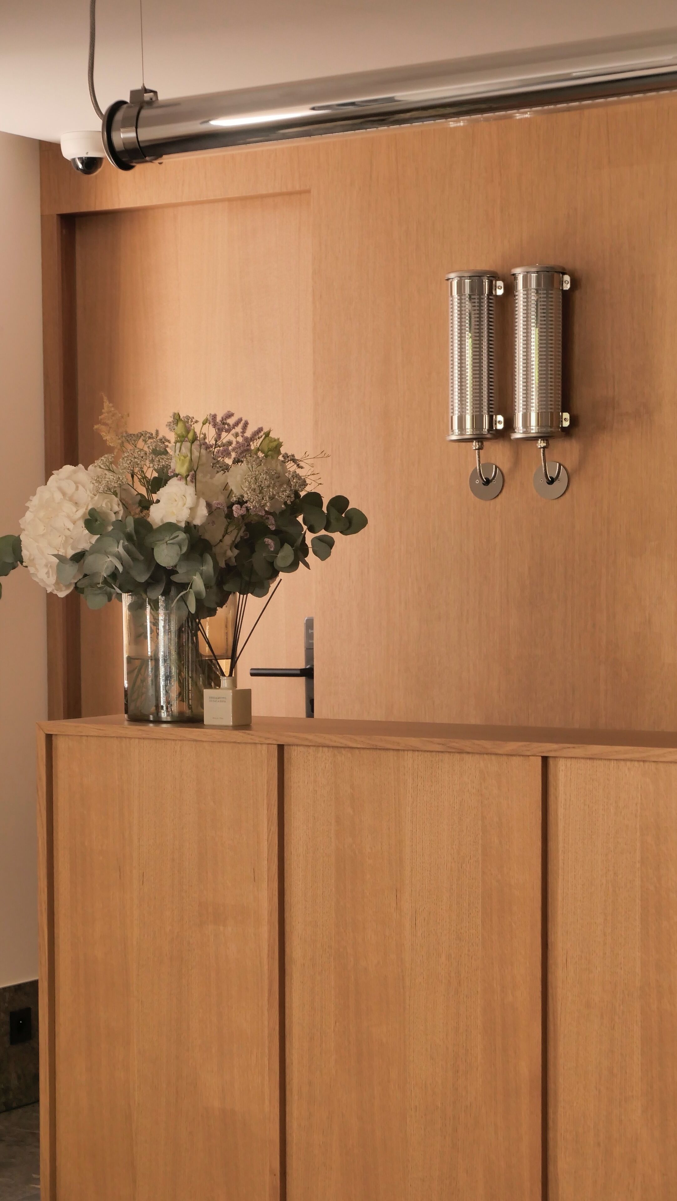 A wooden reception desk with a vase of flowers, a wall-mounted light fixture, and a security camera.