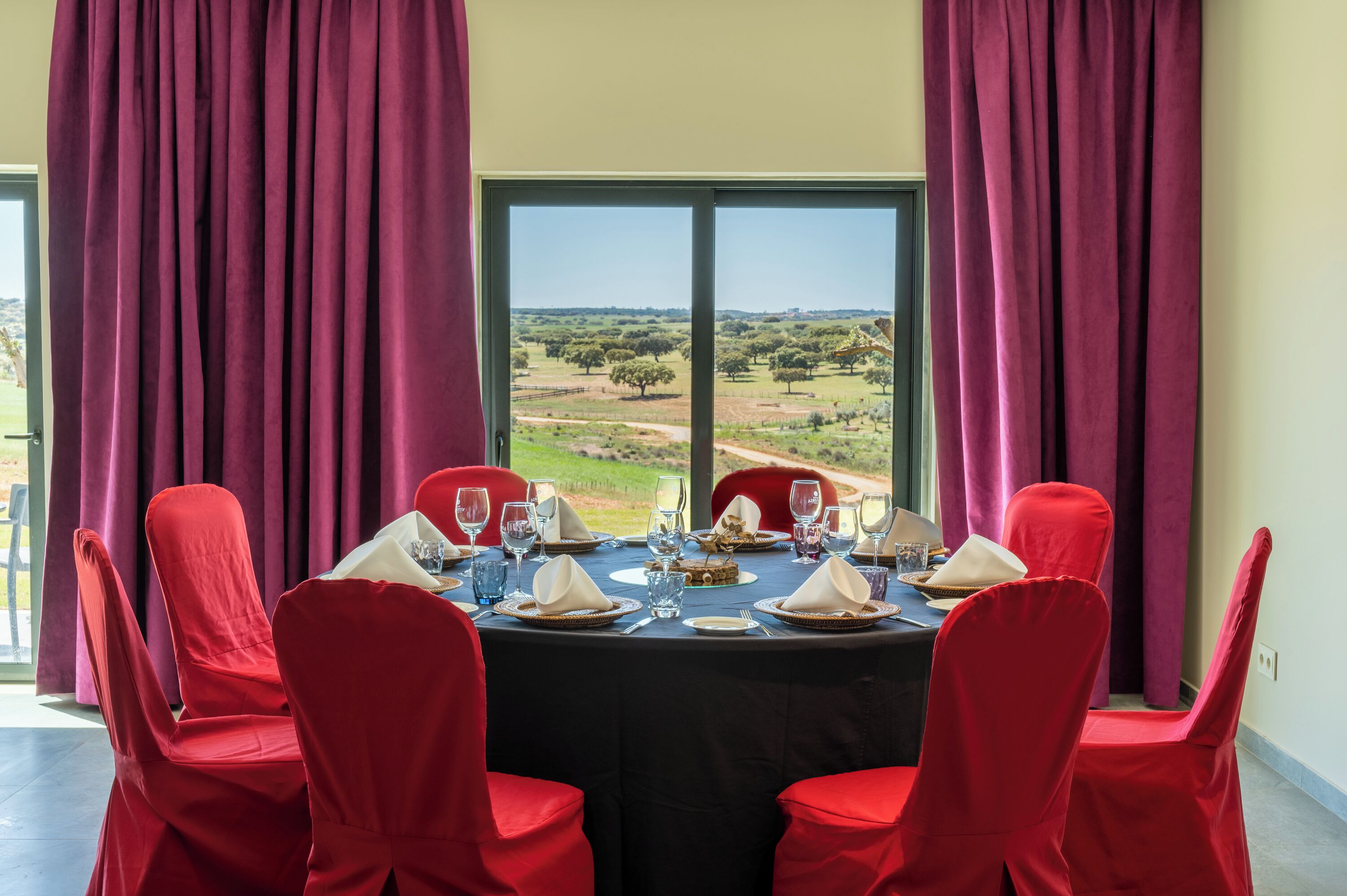 A dining table set for six with red chairs, a view of a landscape through a large window, and a tablecloth.