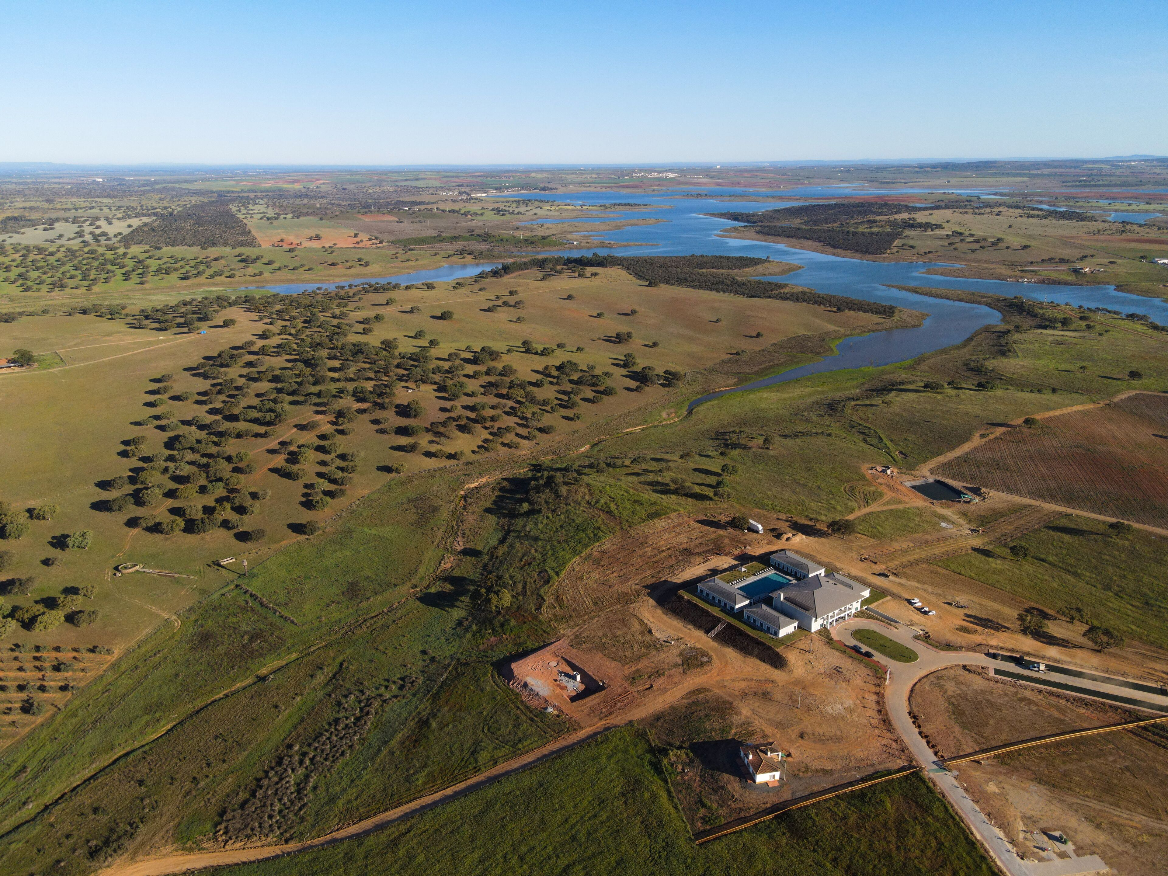 An aerial view of a rural landscape with a large body of water, scattered trees, and a few buildings including a house and a larger structure with a blue roof.