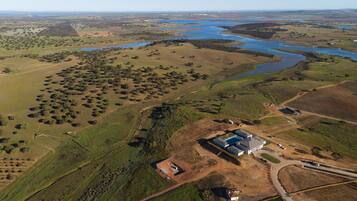 An aerial view of a rural landscape with a large body of water, scattered trees, and a few buildings including a house and a larger structure with a blue roof.