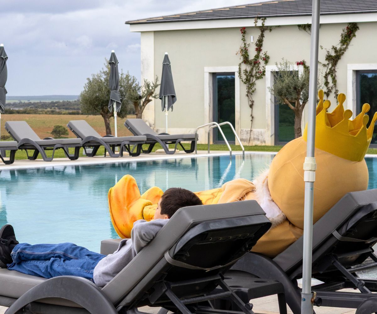 A poolside area with lounge chairs, a person in a yellow costume with a crown, and a building with a window and greenery.
