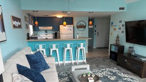 Een woonkamer met een bank, salontafel en barstoelen. In de achtergrond is een keuken met een koelkast en fornuis zichtbaar.