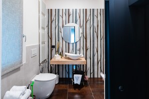 A modern bathroom with a large white toilet, a wooden vanity with a white sink, and a mirror above it. There is a towel rack with towels and a green cleaning brush. The wall has a tree pattern wallpaper. - Sacrati 39 | Tourist Apartment in the Historic Center (Ferrara)