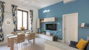 A modern living room with a blue accent wall, a flat-screen TV, a dining area with a table and chairs, and a sofa with a yellow pillow. - OurJoy Condo, 55sm, free private parking and breakfast, high-speed WIFi and A/C (Milano)