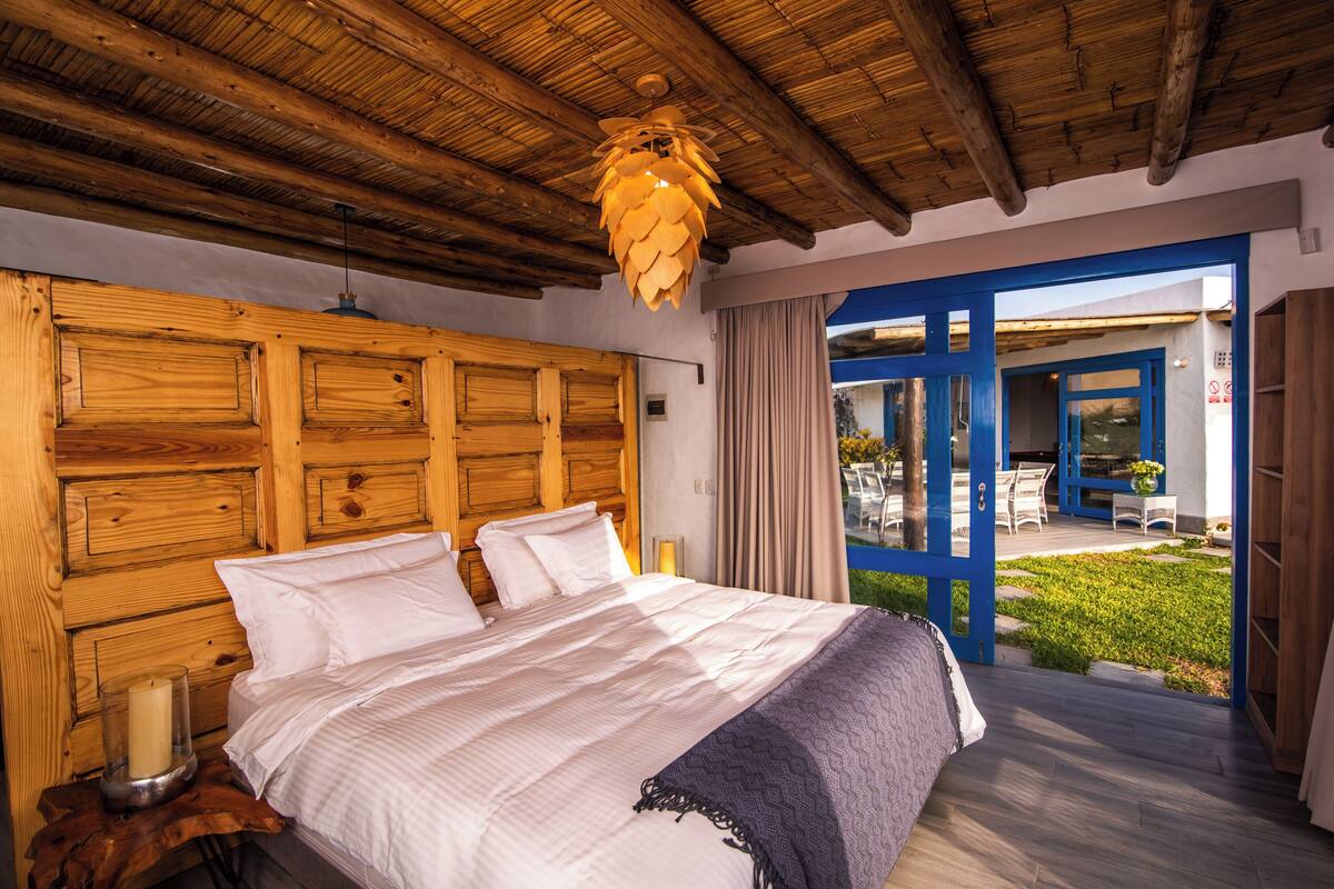 A bedroom with a wooden headboard, a large bed with white linens, a bedside table with a candle, and a view of an outdoor patio with blue doors.