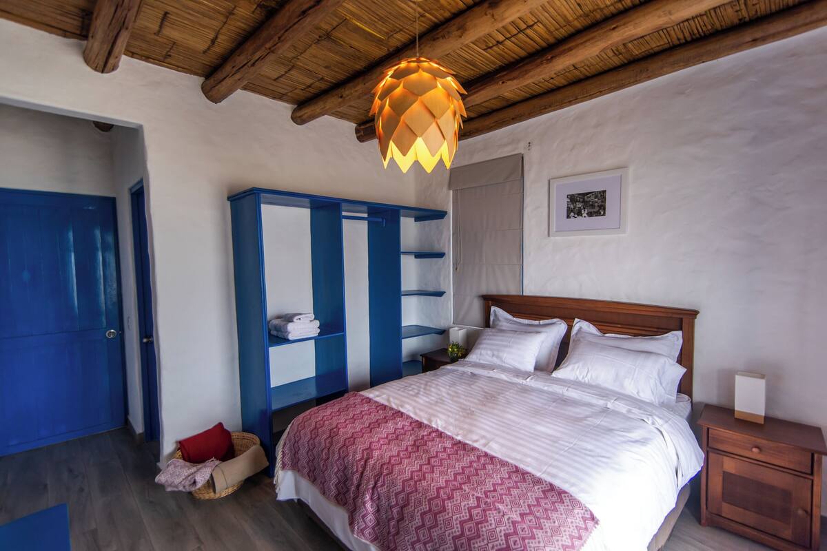 A bedroom with a wooden ceiling, a bed with white bedding, a blue wardrobe, and a small bedside table.