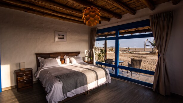 A bedroom with a large bed, wooden ceiling beams, a window with a view of the beach, and a nightstand with a lamp.
