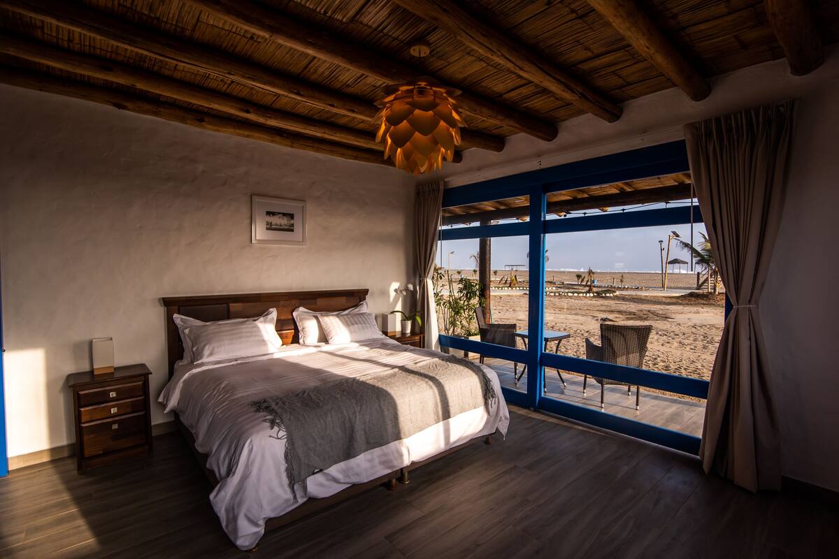 A bedroom with a large bed, wooden ceiling beams, a window with a view of the beach, and a nightstand with a lamp.