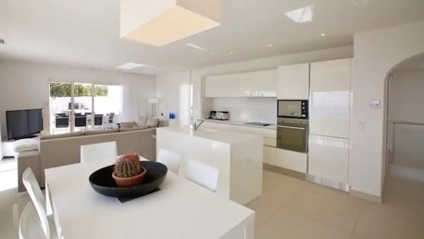 Eine moderne Küche mit weißen Schränken, ein Essbereich mit weißem Tisch und Stühlen sowie ein Wohnbereich mit Sofa und Fernseher.