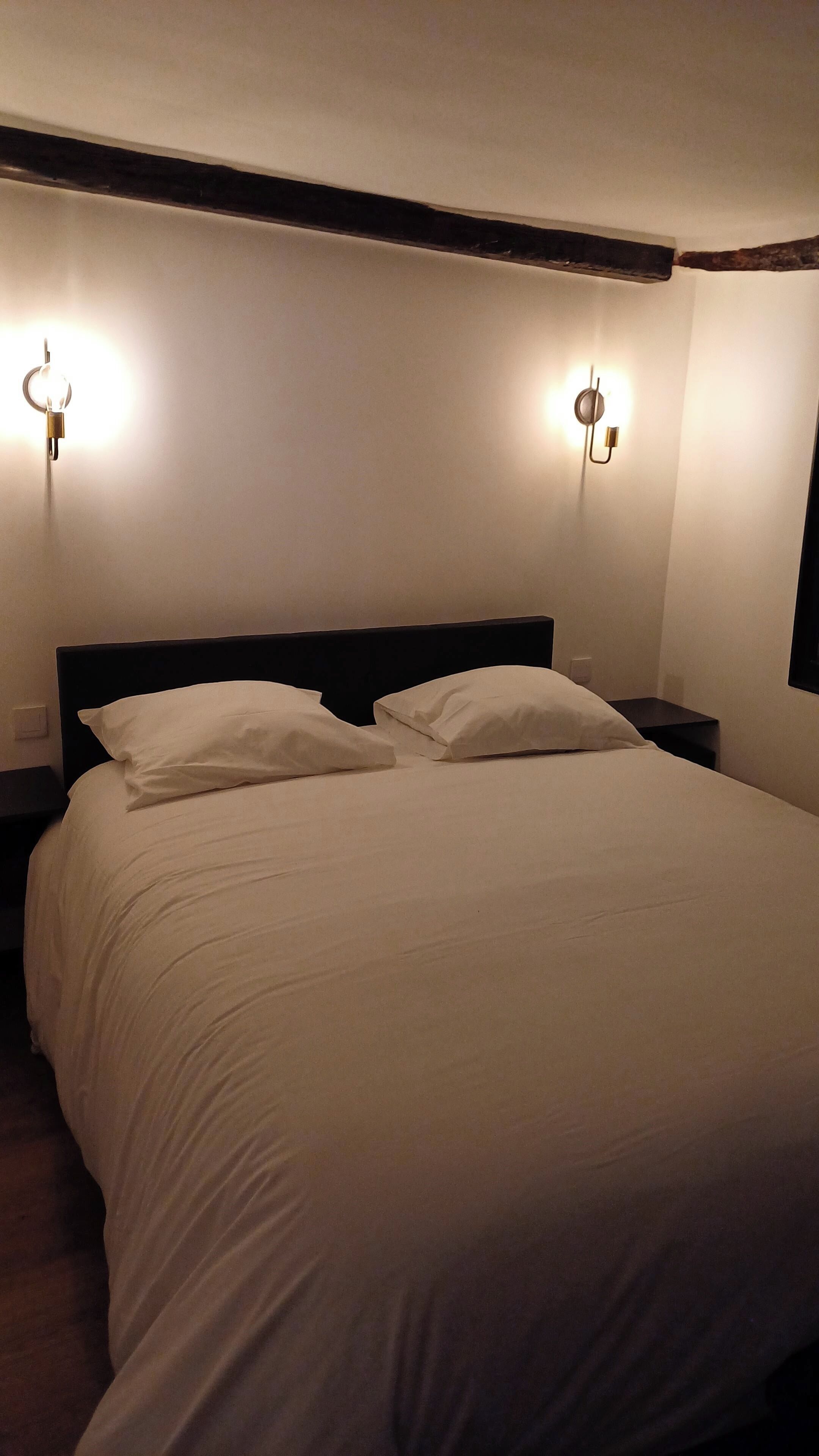 Una cama con ropa de cama blanca, dos lámparas fijadas a la pared y un cabecero oscuro.