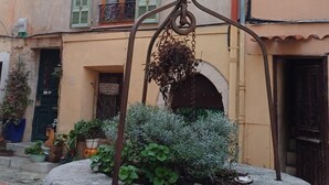 Ein Stebrunnen mit Pflanzen, ein Gebäude mit grünen Fensterläden und eine Metallkonstruktion im Vordergrund.