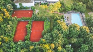 Vista aérea de un complejo con dos canchas de tenis rodeadas de abundante vegetación, una piscina y un edificio de techo plano.