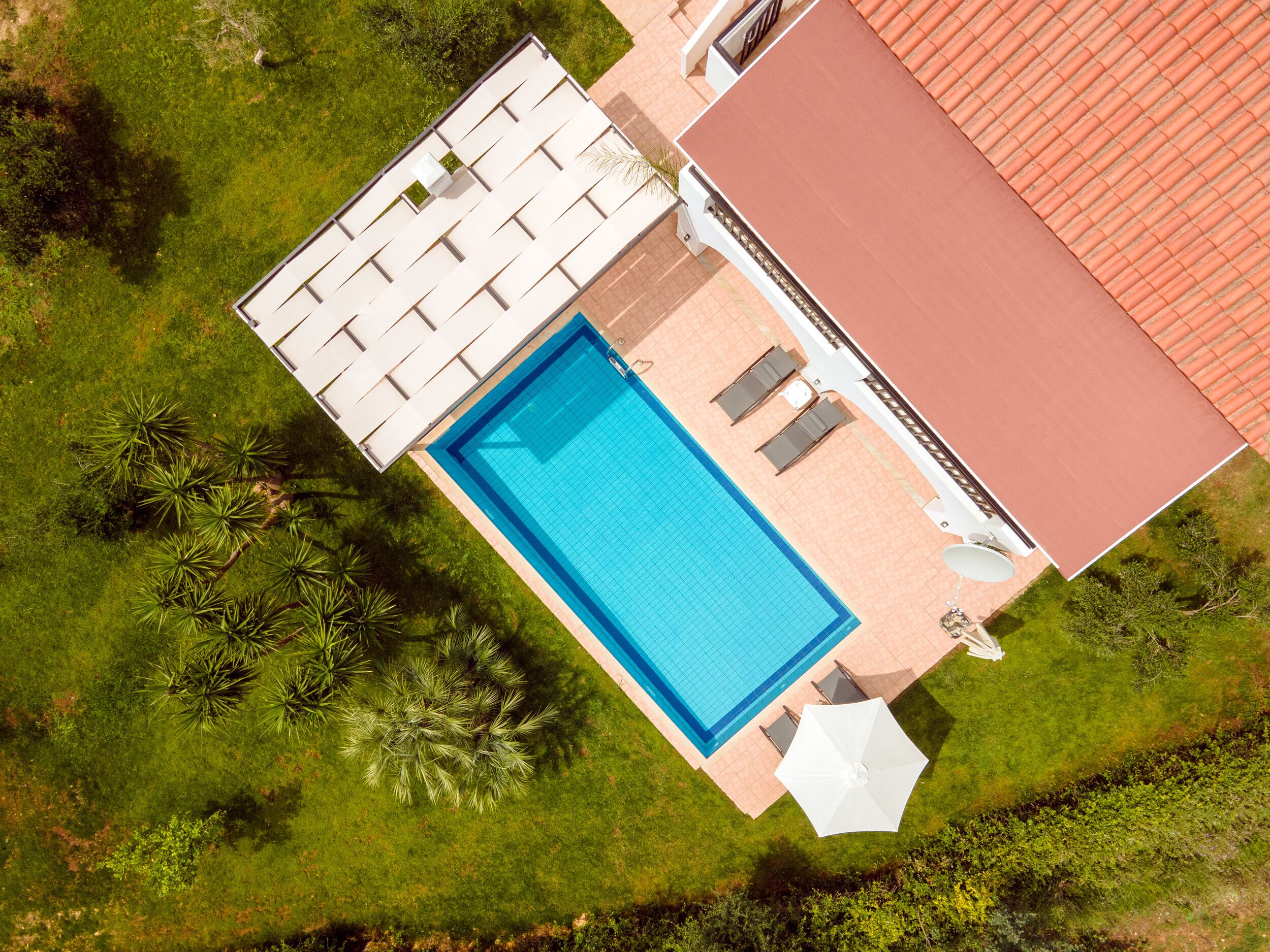 Eine Vogelperspektive eines Schwimmbeckens mit einer gefliesten Terrasse, umgeben von Grünpflanzen und einem Gebäude mit rotem Dach.