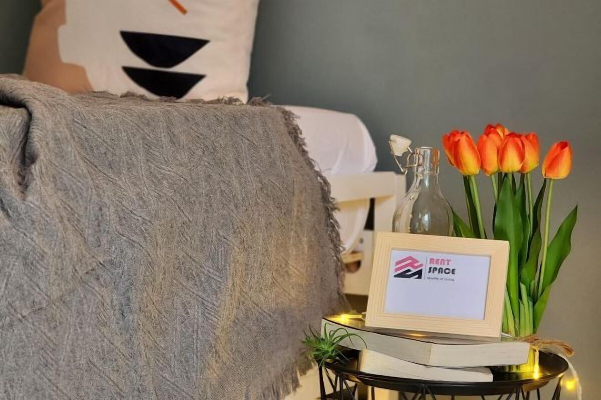 Une pièce chaleureuse avec un canapé gris à la texture intéressante, un panneau encadré indiquant « ESPACE À LOUER », une petite table sur laquelle se trouvent un livre et une bouteille en verre, et un vase rempli de tulipes orange.
