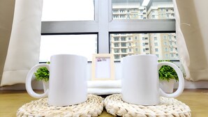 Two white mugs on woven coasters with green plants, placed on a wooden table by a window with a view of buildings. - Ixora Coliving Lifestyle Homestay (Malacca City)