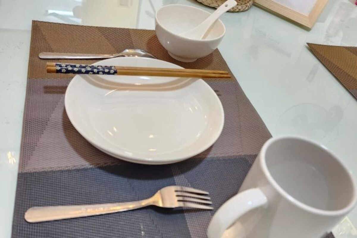 Una preparación para comer con un plato blanco, palillos, un bol blanco, una cuchara blanca, una taza blanca y un tenedor sobre un mantel individual.