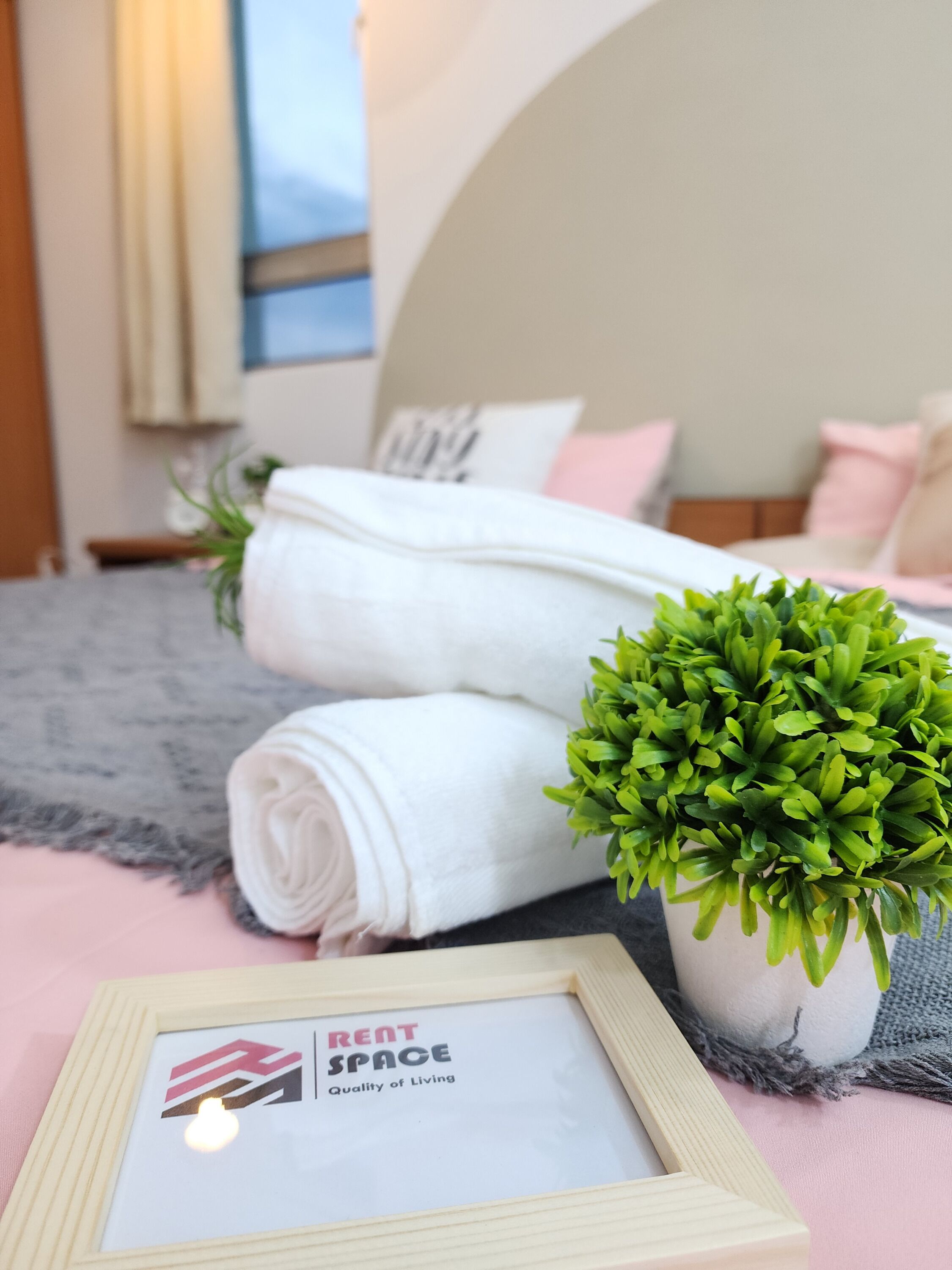 Un lit bien fait, agrémenté de serviettes blanches roulées, d’une petite plante verte et d’une affiche encadrée portant l’inscription : « ESPACE À LOUER – Qualité de vie ».