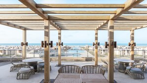 A covered outdoor seating area with tables and wicker chairs, overlooking a cityscape and a body of water. - Gewan Resort New Alamein (Marsa Matrouh)