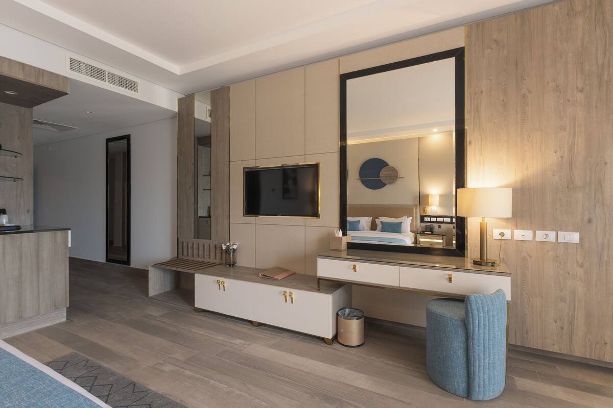 A modern hotel room with a large mirror, a flat-screen TV, a bedside table with a lamp, and a built-in bench.