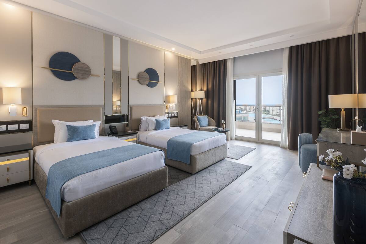 A modern hotel room with two beds, a desk, a chair, and a large window offering a view of the city.