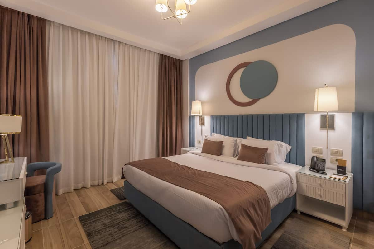 A modern hotel room with a large bed, a desk, and a chair. There is a nightstand with a lamp and a phone.