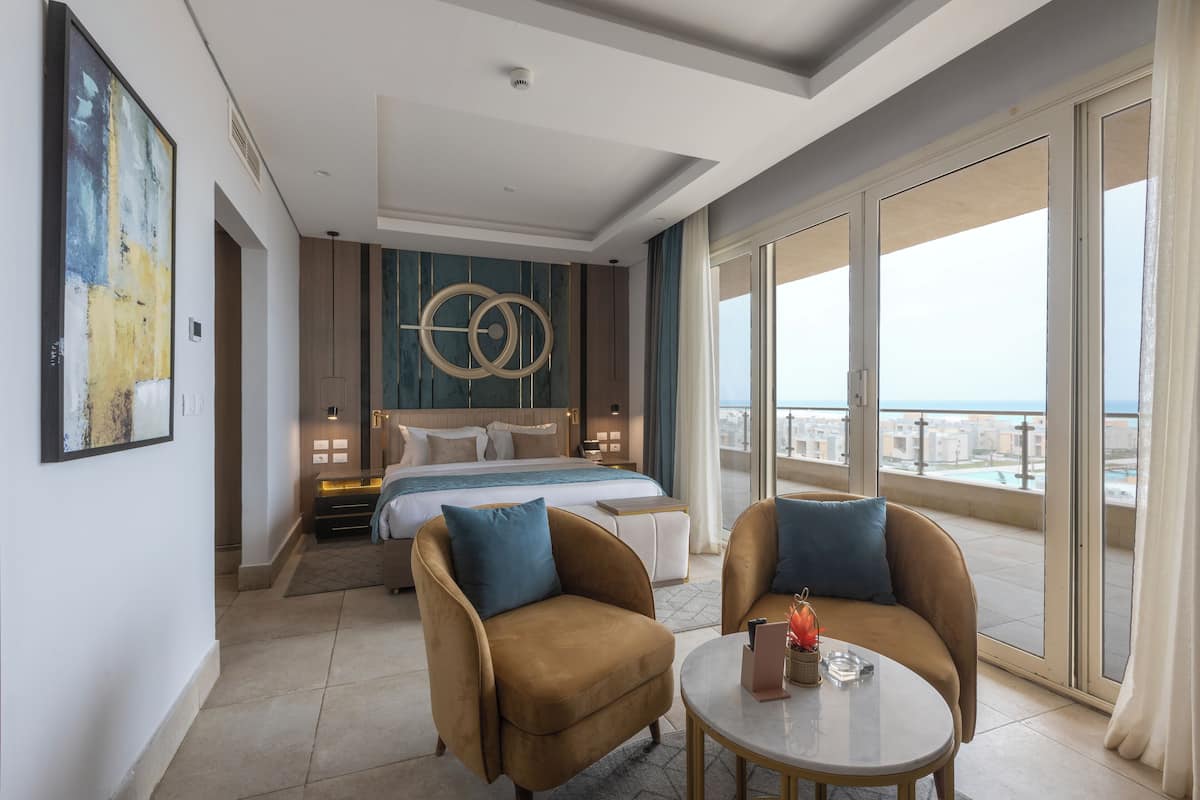 A modern hotel room with a large bed, two armchairs, a small table, and a balcony with a city view.
