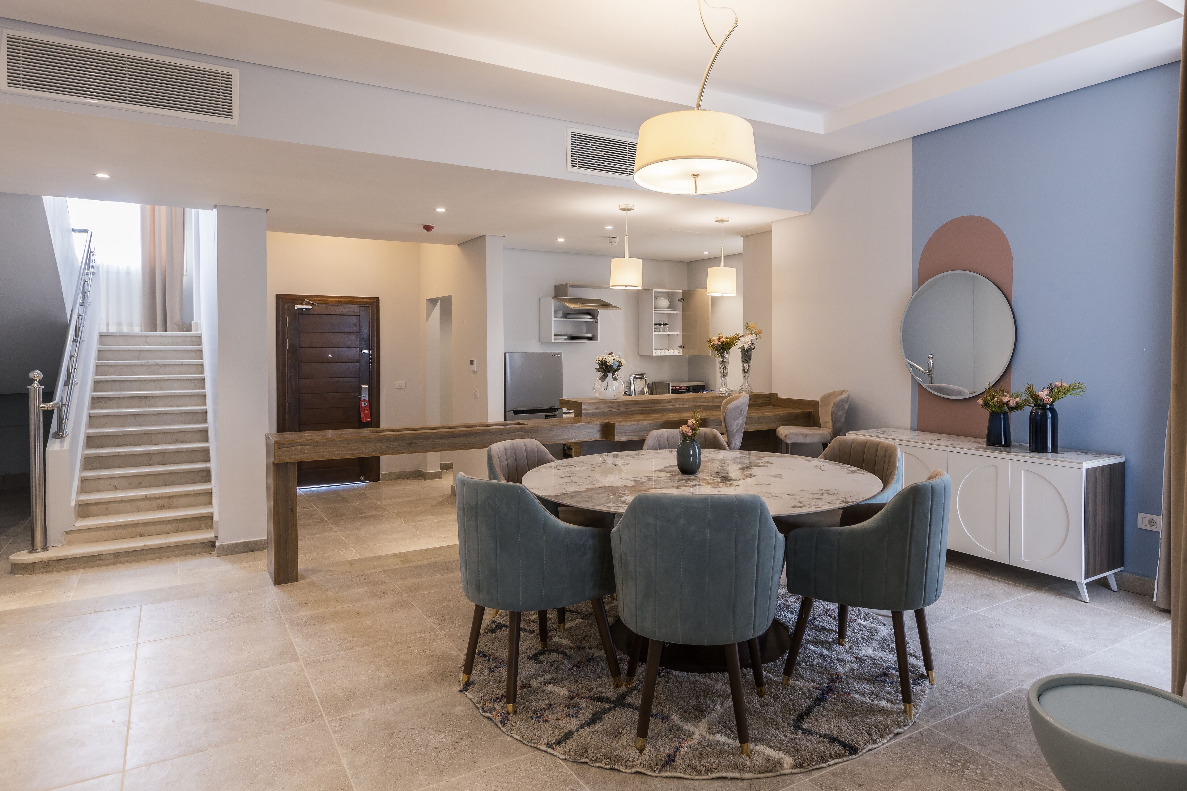 A modern kitchen with a central island, a dining area with a round table and chairs, and a staircase leading to another level.