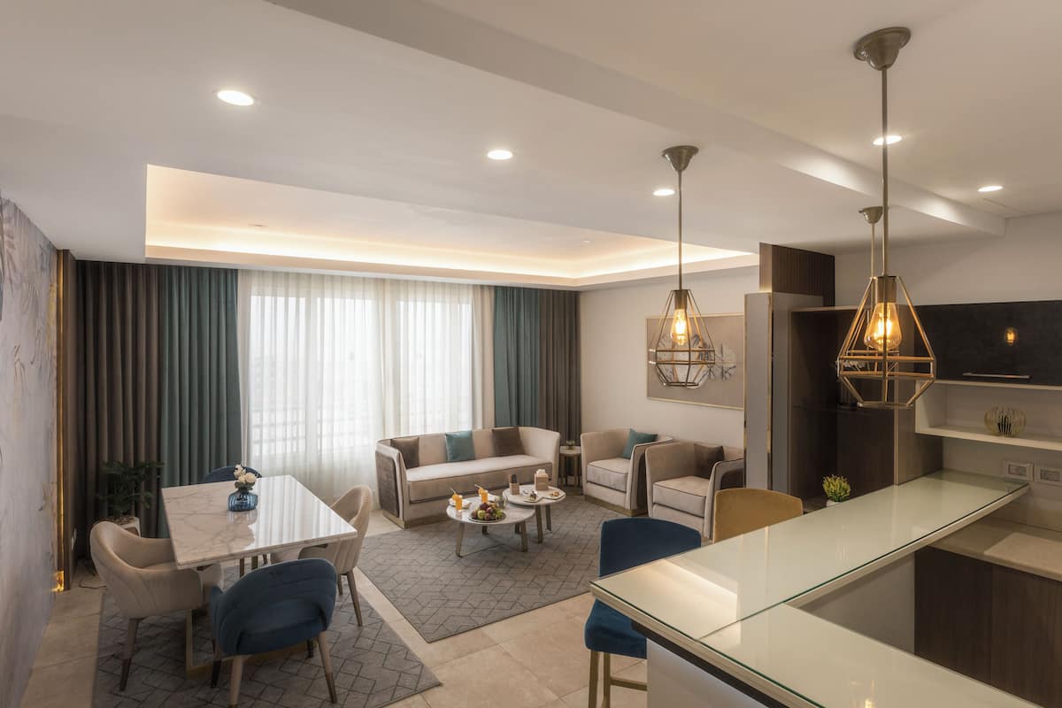 A modern living room with a sofa, coffee table, and dining area. There is a kitchen with a glass island and pendant lighting.
