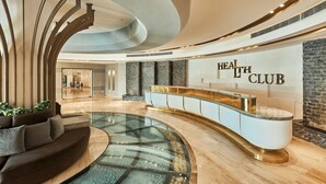 A modern hotel lobby with a curved reception desk, a seating area with a sofa, and a large wall displaying the hotel's name. - Gewan Resort New Alamein (Marsa Matrouh)