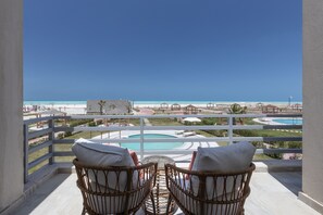 A balcony with wicker chairs and a table, overlooking a pool and beach area.