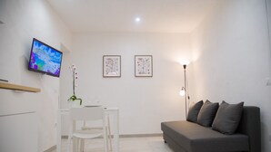 Una sala moderna con un sofá gris, una mesa de comedor blanca con sillas, un televisor montado en la pared y dos cuadros enmarcados.