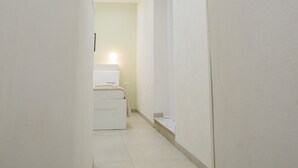 Un pasillo estrecho con una puerta blanca al final, que da a una habitación con una cama y una mesita de noche.