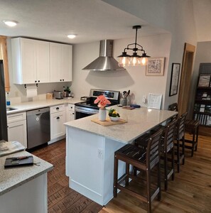 Una cocina moderna con armarios blancos, extractor de acero inoxidable, una isla central con taburetes de bar y una lámpara colgante.