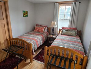 Una habitación con dos camas individuales, una silla de madera, una mesita de noche, una lámpara y un ventanal con cortinas.