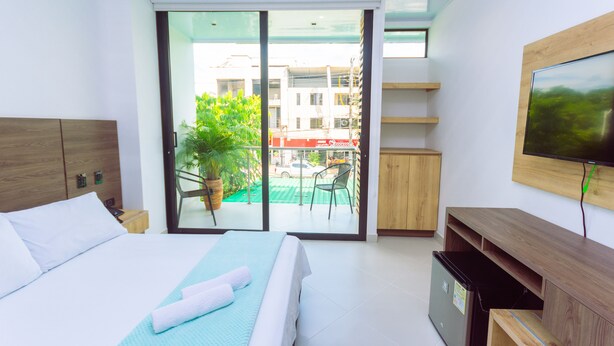 Una habitación de hotel moderna con una cama, un televisor, un ventanal corredero que da a un balcón y una pequeña cocina.