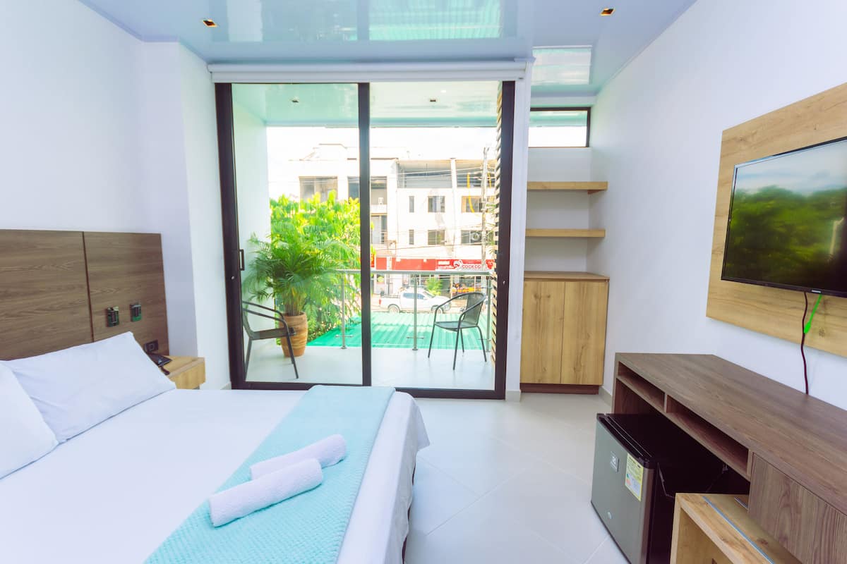 A modern hotel room with a bed, a TV, a sliding glass door leading to a balcony, and a small kitchenette.