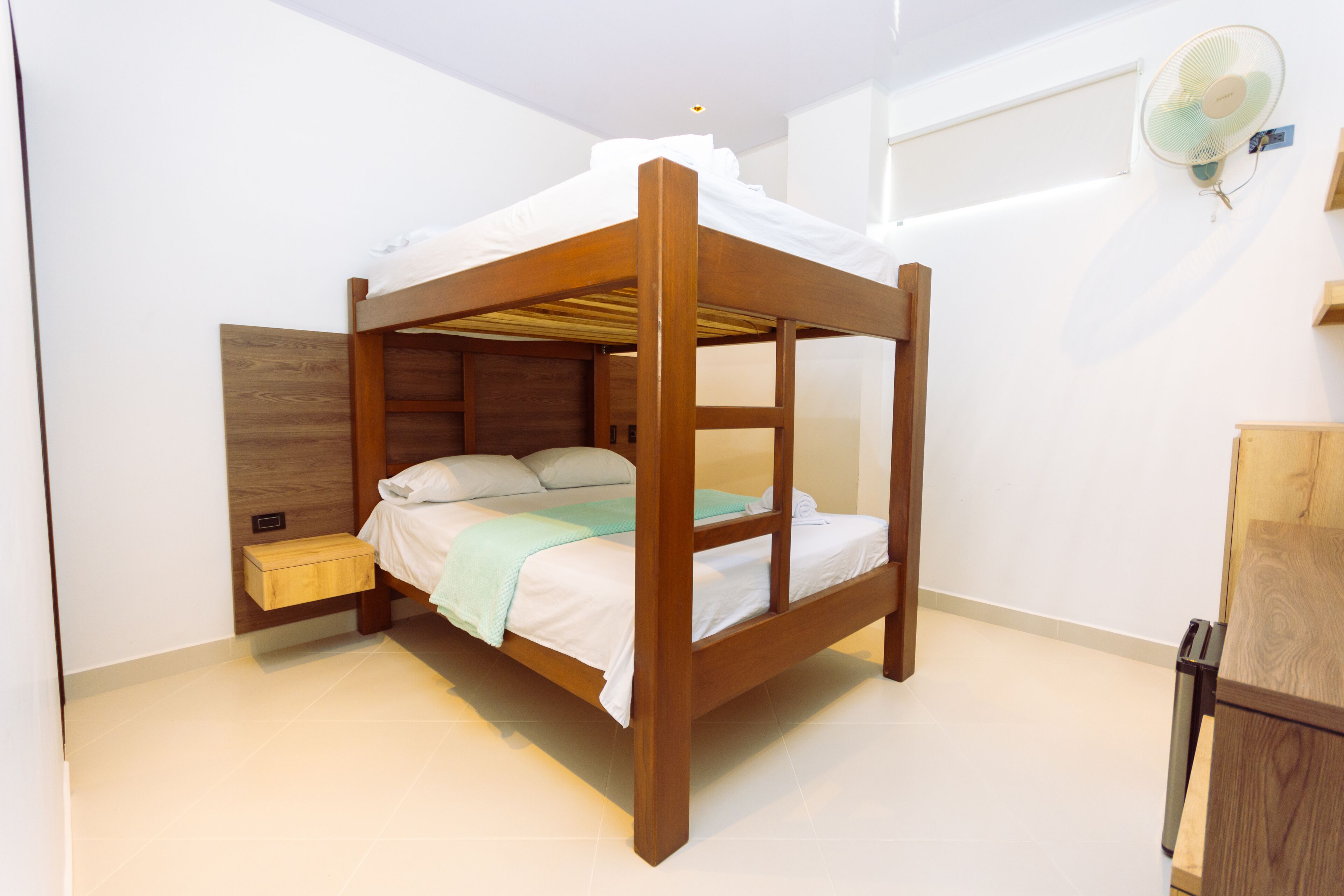 A single bed with a wooden frame and white bedding, a bedside table, and a wall-mounted fan.