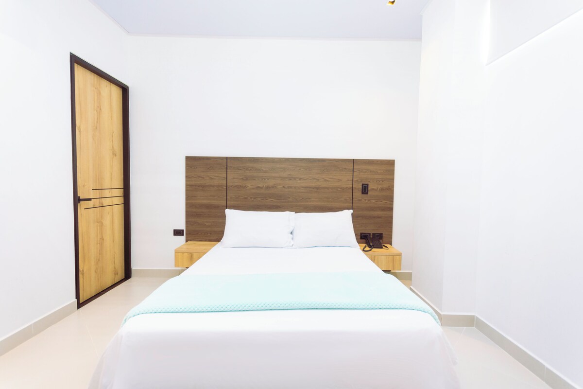 A modern hotel room with a wooden headboard, a bed with white linens, a nightstand, and a wooden door.