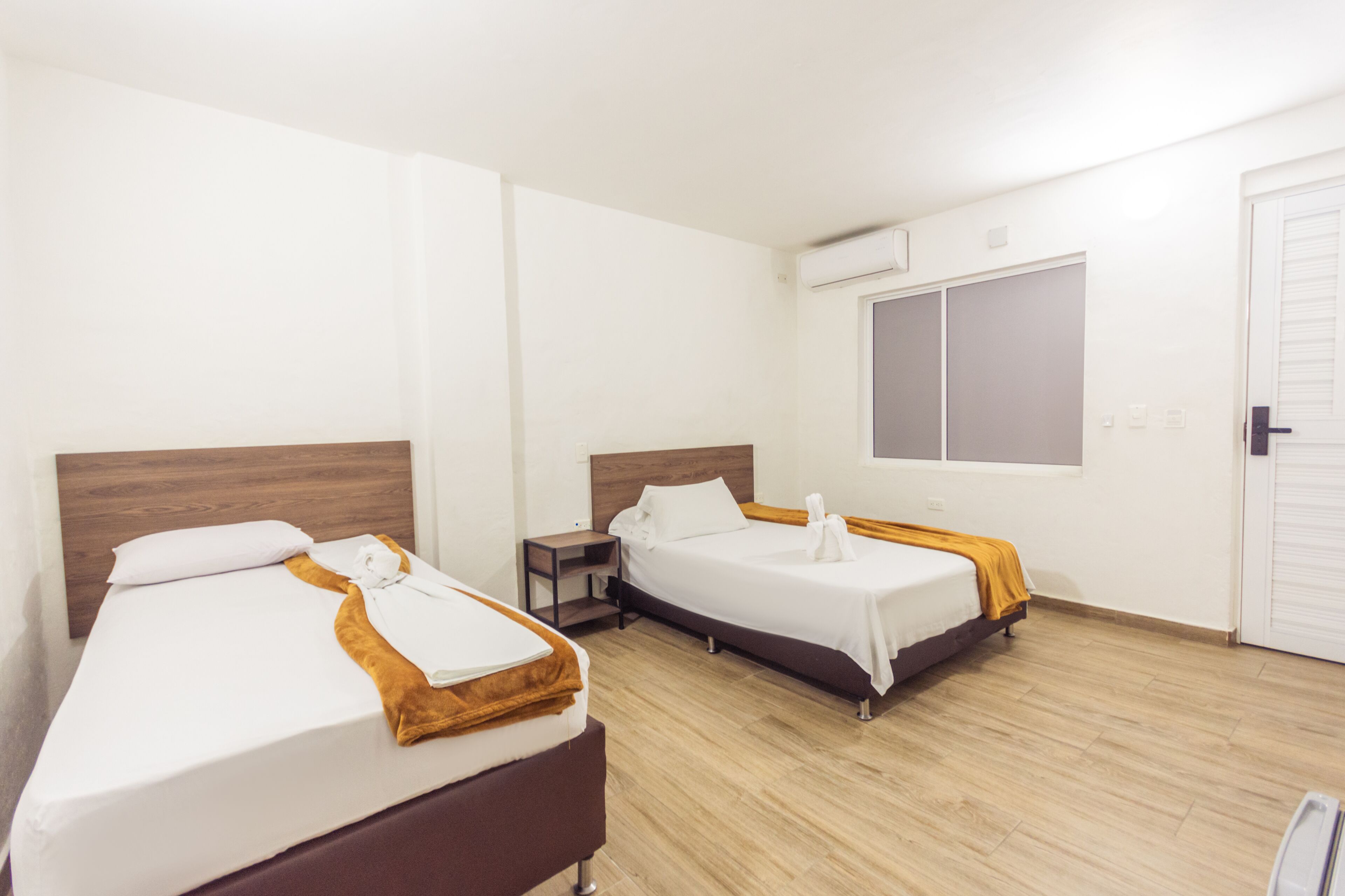 A hotel room with two beds, a wooden headboard, a small bedside table, and a window with blinds.