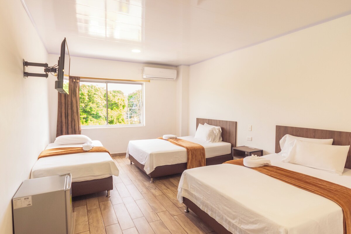 A hotel room with three beds, a TV, an air conditioner, and a window with a view of greenery.