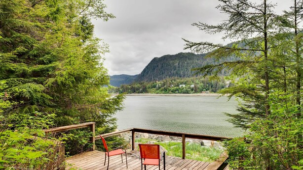A wooden deck with two chairs overlooking a lake, surrounded by dense forest and mountains. - Juneau Vacation Home: Stunning View + Beach Access (Juneau)