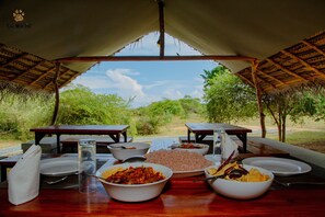 An outdoor dining area with a view of the landscape, tables with plates of food, and a thatched roof structure. - Yala Wild Hut (Thissamaharama)